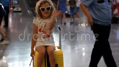 小女孩游客坐在火车站的一个黄色的大行李箱上，挥手告别。 孩子正在等待